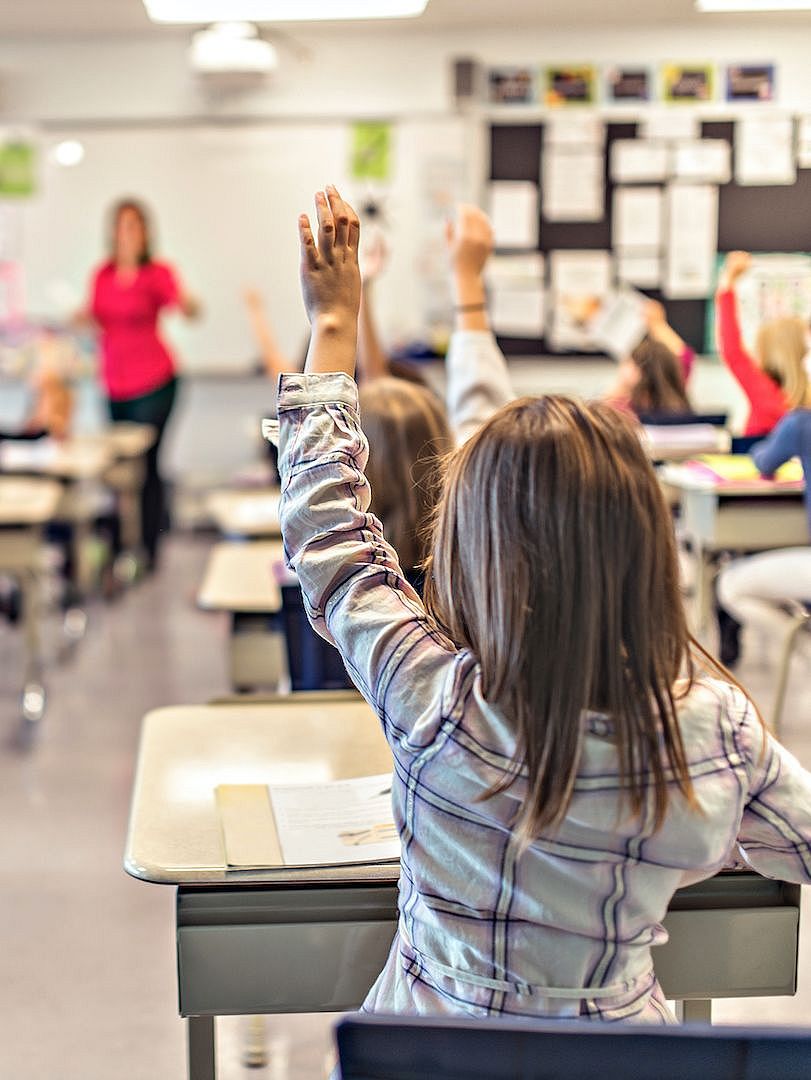 Bild von Klassenraum von hinten, Kind mit braunen langen Haaren hebt die Hand